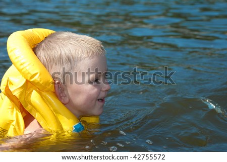 Smiling boy in a yellow life jacket is swimming in the river