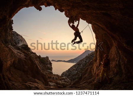 Male rock climber hanging on challenging route on cliff at sunset, female climber belaying him