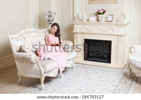 Young girl in elegant pink dress reading book while sitting on couch