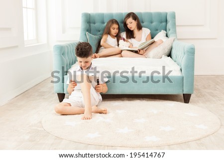 Siblings spending free time at home: two girls reading book on couch and boy using tablet while sitting on floor