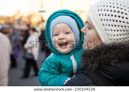 Joyful baby boy and his mother outdoors in wintertime, with blurred people in background