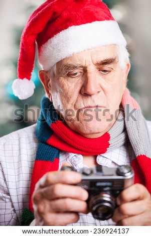 Senior man in Santa\'s hat looking at display of retro style camera on Christmas background