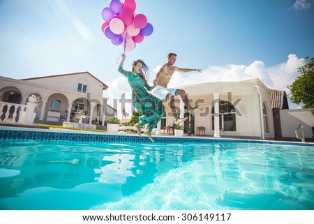 Happy young couple jumping into the pool while holding a bunch of balloons