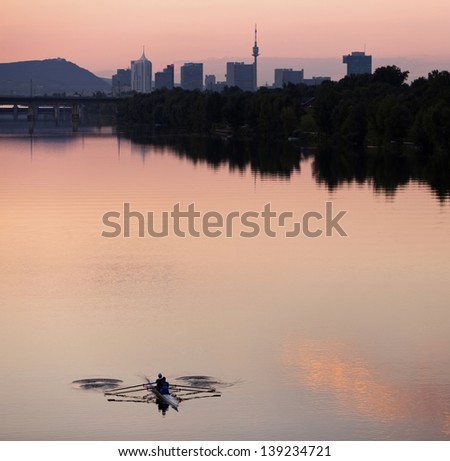 Rowing guys at dusk in the Danube River at Vienna - Austria with urban skyline of the Danube City, UNO City, Donauturm, Leopoldsberg, etc. in the background.