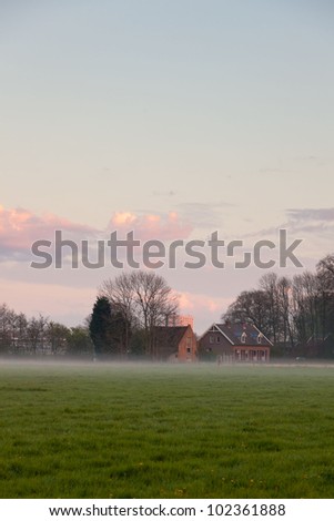 Meadow with house in the mist at sunset. Cloudy sky. Spring time. City skyline.