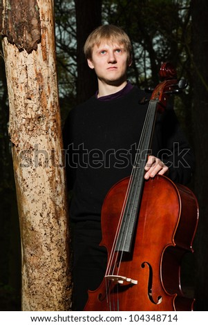 Portrait of young male cello player in green spring forest. Blond hair. Playing classic instrument. Dressed in black. Artistic looking.