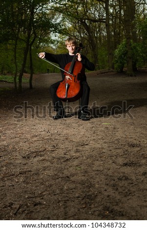 Portrait of young male cello player in green spring forest. Blond hair. Playing classic instrument. Dressed in black. Artistic looking.