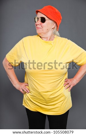 Funny and happy senior woman wearing yellow shirt and orange hat and sunglasses. Cool and hip. Studio shot isolated on grey.