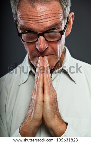 Expressive good looking senior man with glasses against grey wall. Hands praying. Spiritual and characteristic. Well dressed. Studio shot.