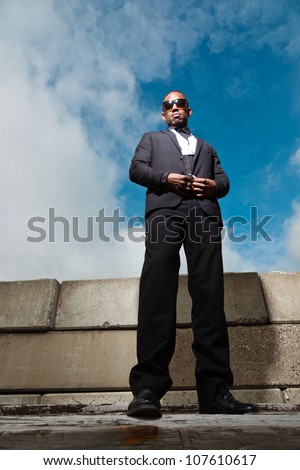 Cool black american man in dark suit wearing sunglasses. Fashion shot in urban setting. Blue cloudy sky.