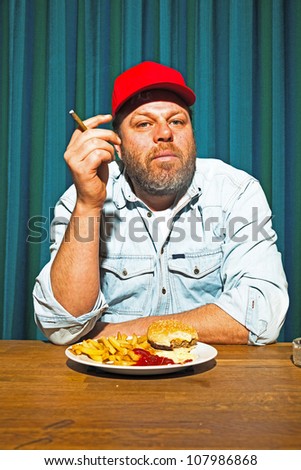 Man with beard eating fast food meal. Enjoying french fries and a hamburger. Smoking a cigar. Trucker with red cap.
