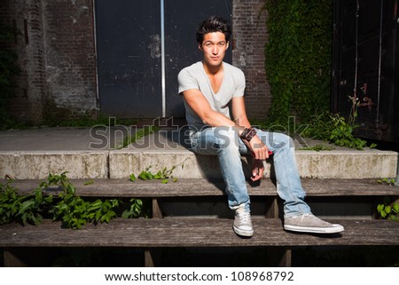 Urban asian man sitting on stairs. Good looking. Cool guy. Wearing grey shirt and jeans. Old neglected building in the background.