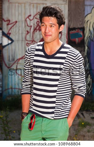 Urban asian man. Good looking. Cool guy. Wearing blue white striped sweater and green shorts. Standing in front of wooden wall with graffiti.