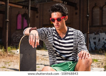 Urban asian man with red sunglasses and skateboard sitting on street. Good looking. Cool guy. Wearing blue white striped sweater and green shorts. Old neglected building in the background.