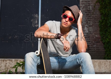 Urban asian man with hat, red sunglasses and skateboard sitting on stairs. Good looking. Cool guy. Wearing grey shirt and jeans. Old neglected building in the background.