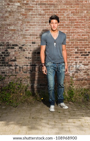 Urban asian man. Good looking. Cool guy. Wearing grey shirt and jeans. Standing in front of brick wall.