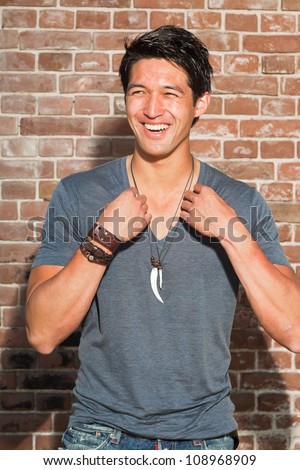 Smiling urban asian man. Good looking. Cool guy. Wearing grey shirt. Standing in front of brick wall.