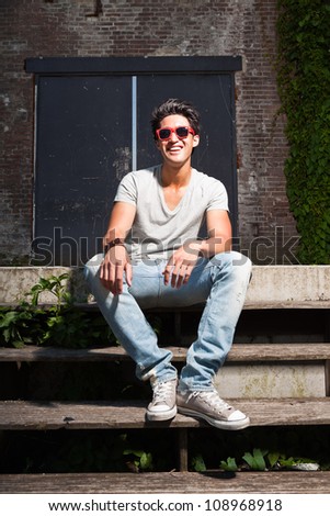 Urban asian man with red sunglasses sitting on stairs. Good looking. Cool guy. Wearing grey shirt and jeans. Old neglected building in the background.