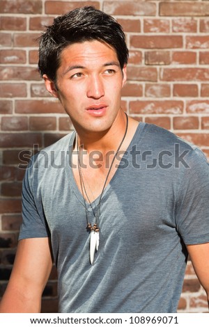 Urban asian man. Good looking. Cool guy. Wearing grey shirt. Standing in front of brick wall.