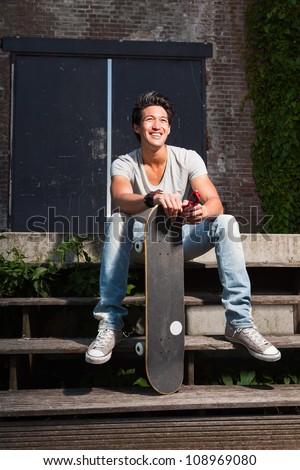 Urban asian man with skateboard sitting on stairs. Good looking. Cool guy. Wearing grey shirt and jeans. Old neglected building in the background.