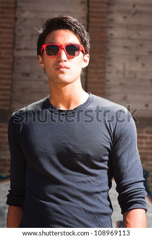 Urban asian man with red sunglasses. Good looking. Cool guy. Wearing dark blue shirt. Standing in front of brick wall with graffiti.