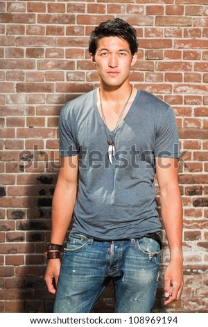 Urban asian man. Good looking. Cool guy. Wearing grey shirt and jeans. Standing in front of brick wall.