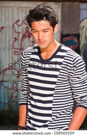 Urban asian man. Good looking. Cool guy. Wearing blue white striped sweater. Standing in front of wooden wall with graffiti.