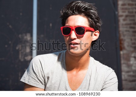 Urban asian man with red sunglasses. Good looking. Cool guy. Wearing grey shirt and jeans. Old neglected building in the background.