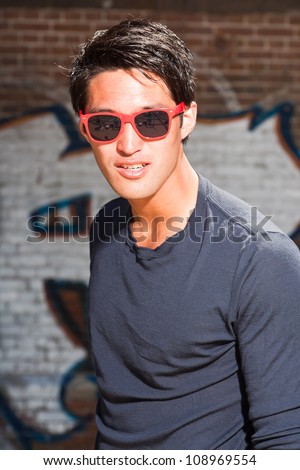 Urban asian man with red sunglasses. Good looking. Cool guy. Wearing dark blue shirt. Standing in front of brick wall with graffiti.
