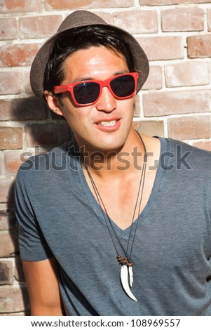 Urban asian man with red sunglasses. Good looking. Cool guy. Wearing grey shirt and hat. Standing in front of brick wall.