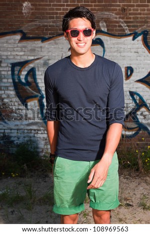 Urban asian man with red sunglasses. Good looking. Cool guy. Wearing dark blue shirt and green shorts. Standing in front of brick wall with graffiti.