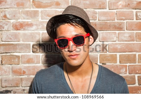Urban asian man with red sunglasses. Good looking. Cool guy. Wearing grey shirt and hat. Standing in front of brick wall.