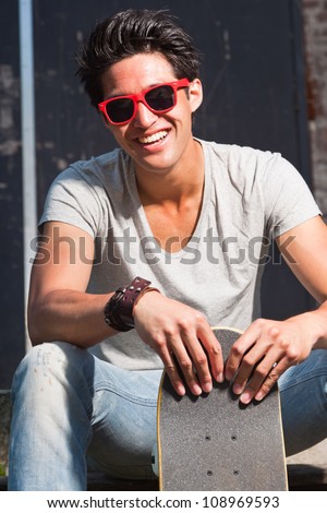 Urban asian man with red sunglasses and skateboard sitting on stairs. Good looking. Cool guy. Wearing grey shirt and jeans.