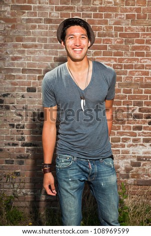 Urban asian man. Good looking. Cool guy. Wearing grey shirt and hat and jeans. Standing in front of brick wall.