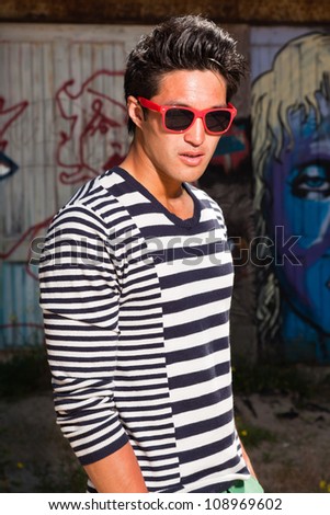 Urban asian man with red sunglasses. Good looking. Cool guy. Wearing blue white striped sweater. Standing in front of wooden wall with graffiti.