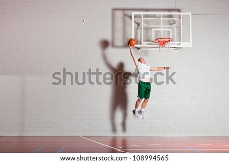Tough healthy young man playing basketball in gym indoor. Wearing white shirt and green shorts.