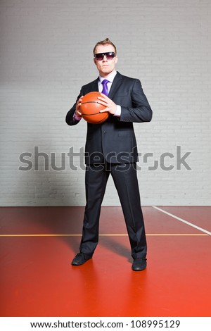 Business man with basketball. Wearing dark sunglasses. Good looking young man with short blond hair. Gym indoor.