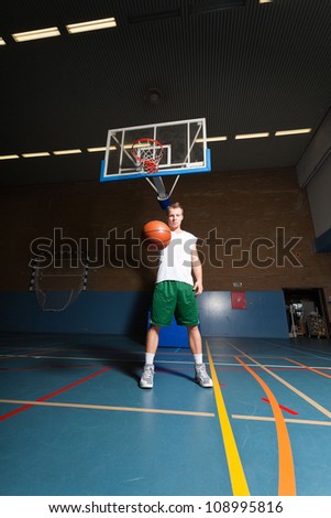 Tough healthy young man playing basketball in gym indoor. Wearing white shirt and green shorts.