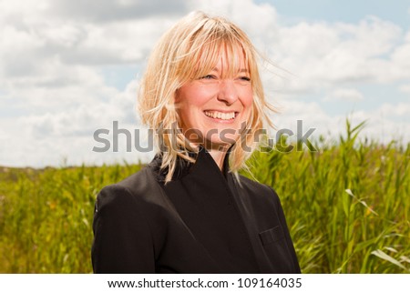 Happy pretty young blond woman enjoying nature. Field with high grass. Blue cloudy sky. Black jacket.
