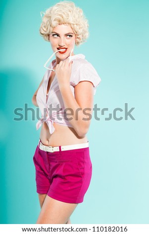 Sexy blonde pin up girl wearing pink shirt and hot pants holding sunglasses. Retro style. Fashion studio shot isolated on light blue background.