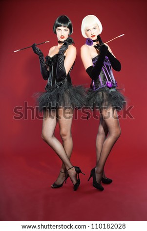 Two burlesque pin up women with black hair dressed in purple and black. Sexy pose. Wearing black hat. Studio fashion shot isolated on red background.
