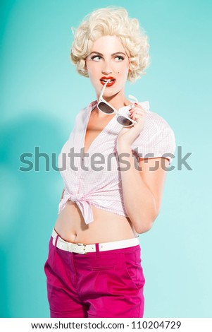 Sexy blonde pin up girl wearing pink shirt and hot pants holding sunglasses. Retro style. Fashion studio shot isolated on light blue background.