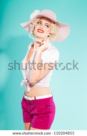 Sexy blonde pin up girl with hat wearing pink shirt and hot pants holding sunglasses. Retro style. Fashion studio shot isolated on light blue background.