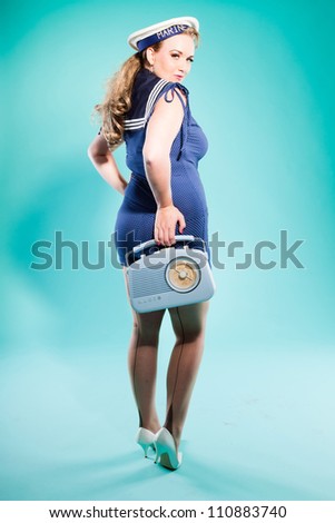 Sexy blonde pin up girl wearing blue dress with white dots and marine cap. Holding vintage radio. Retro style. Fashion studio shot isolated on light blue background.