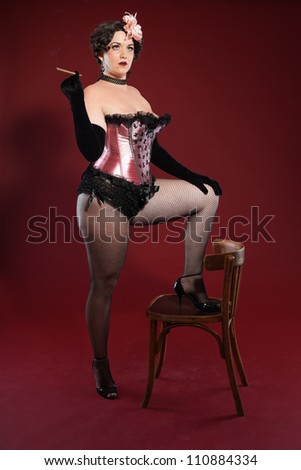 Sexy burlesque pin up woman with long blond hair dressed in pink and black. Smoking cigar. Standing on retro wooden chair. Studio fashion shot isolated on red background.