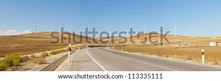 Beautiful panoramic photo of road through agrarian mountain landscape with windmills. On the road. Blue cloudy sky. Golden fields. Amazing scenery. Andalusia. Spain.