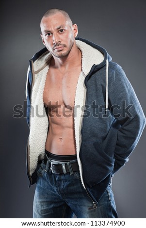 Muscled fitness man. Cool looking. Tough guy. Brown eyes. Bald. Wearing blue hood shirt. Tanned skin. Studio shot isolated on grey background.