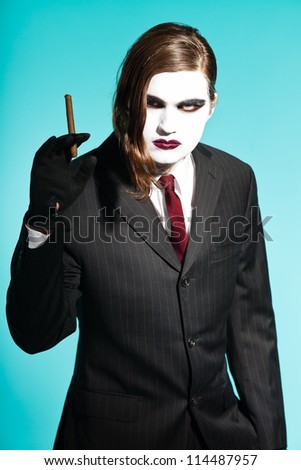 Gothic vampire looking business man wearing black striped suit and dark red tie. Smoking a cigar. Another kind. Scary white face. Isolated on light blue background. Studio shot.