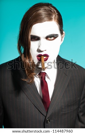 Gothic vampire looking business man wearing black striped suit and dark red tie. Smoking a cigar. Another kind. Scary white face. Isolated on light blue background. Studio shot.