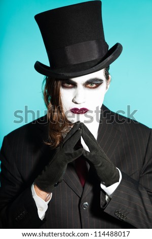 Gothic vampire looking business man wearing black striped suit and dark red tie. Another kind. Scary white face. Black vintage hat. Isolated on light blue background. Studio shot.